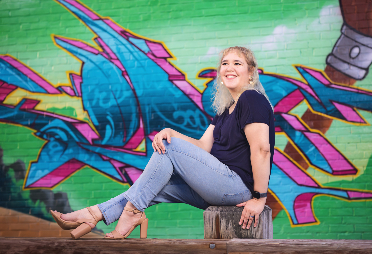 photograph of woman in a navy shirt and light blue jeans sitting on a fence in front of a brick wall covered in graffiti