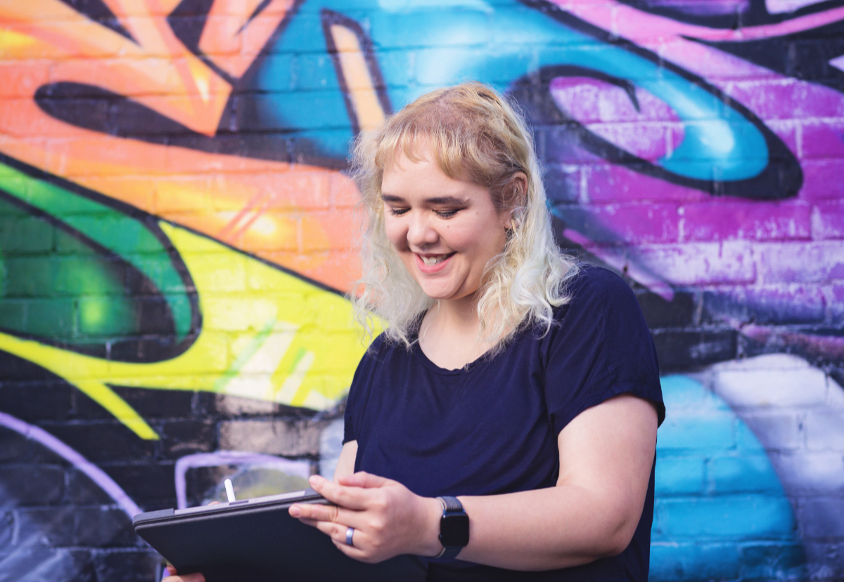 a woman with died silver hair sits in front of a grafitti wall looking down at a drawing tablet in her hands
